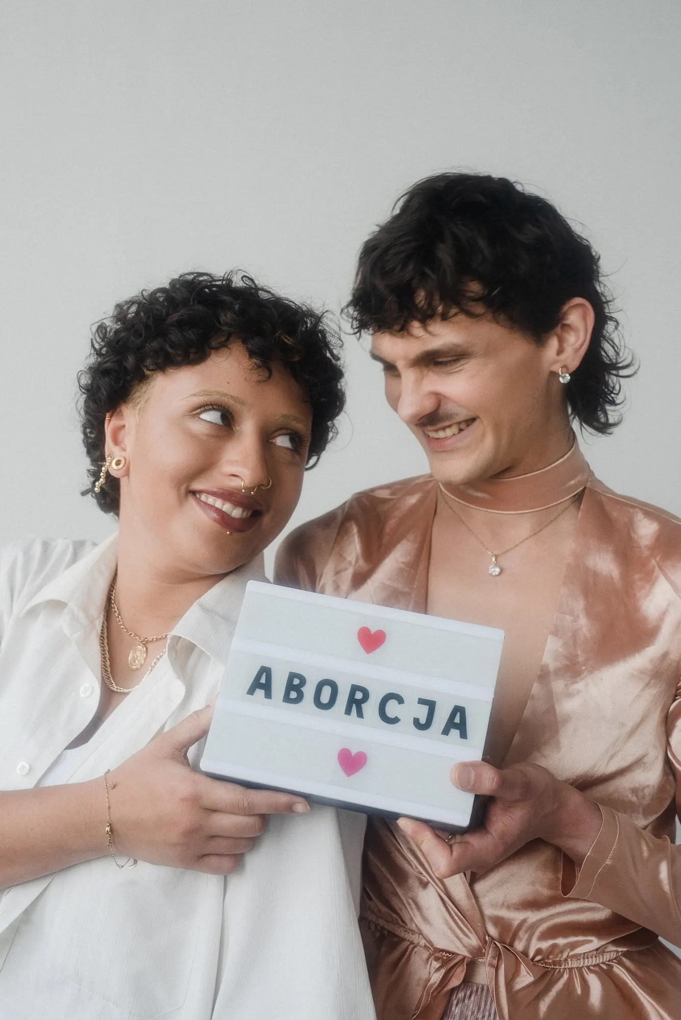 Uśmiechnięta para osób trzymająca mały baner z napisem aborcja. Wokół napisu umieszcozne są małe różowe serduszka.